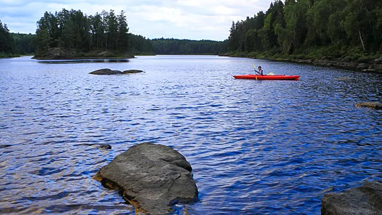 A person kayaking in a red kayak on a tranquil, expansive river surrounded by dense forest, with large rocks emerging from the water's surface.

