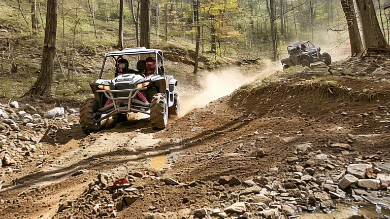 Two UTVs driving through a muddy forest trail, with the lead vehicle splashing through a puddle and the second creating a trail of dust.

