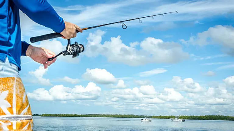 A close-up of a person in a blue shirt and patterned shorts fishing with a rod and reel, set against a backdrop of a serene lake with other boats and a cloudy sky.

