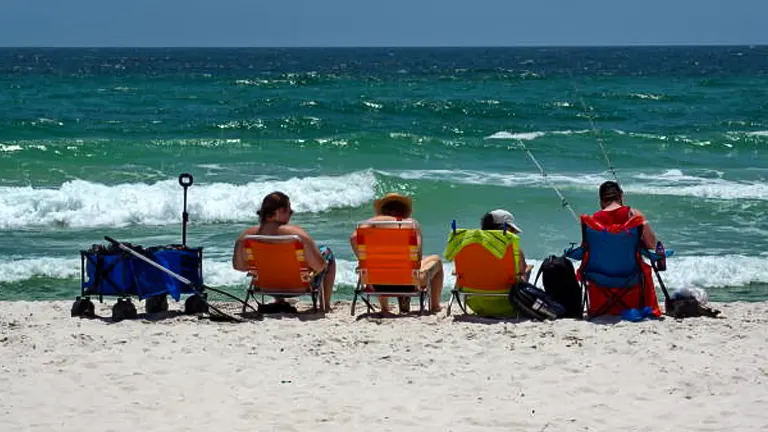 Four individuals seated in colorful beach chairs on the sand facing the sea, with fishing rods set up in front of them, enjoying a bright, sunny day at the beach.
