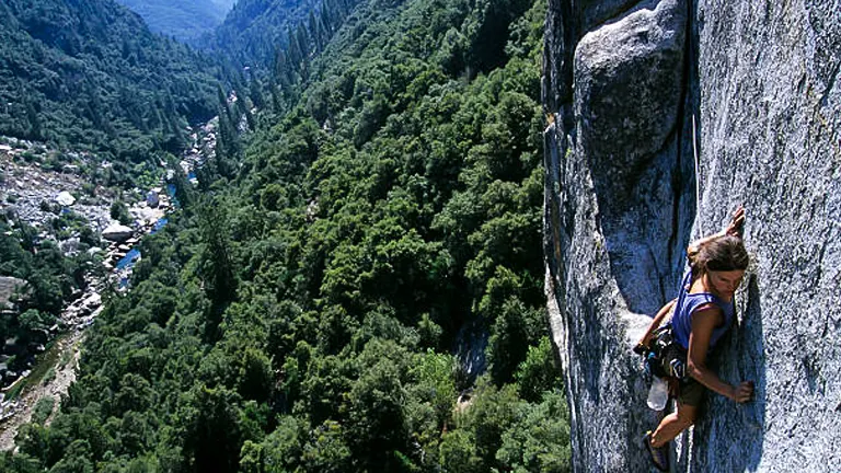 A climber ascending a steep granite cliff with dense forest and a small town visible in the valley below.
