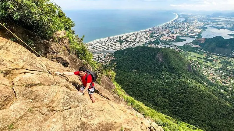 A climber in red and black attire ascending a rocky slope with a panoramic view of a coastal cityscape and lush greenery below.


