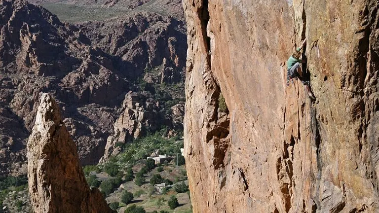 A rock climber in green scaling a steep, rugged cliff with a view of a canyon and a village below.

