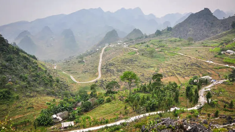 An expansive view of a winding road cutting through a rural, hilly landscape with karst mountain peaks, a patchwork of fields, and scattered houses, under a hazy sky.

