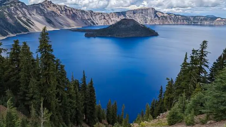 A panoramic view of Crater Lake with the distinctive Wizard Island in the center, surrounded by deep blue water. The lake is framed by steep crater walls and coniferous trees in the foreground under a partly cloudy sky.