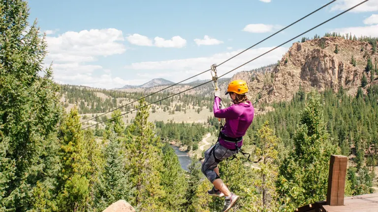 A person in a purple top and helmet ziplining over a forested valley with mountainous terrain in the background