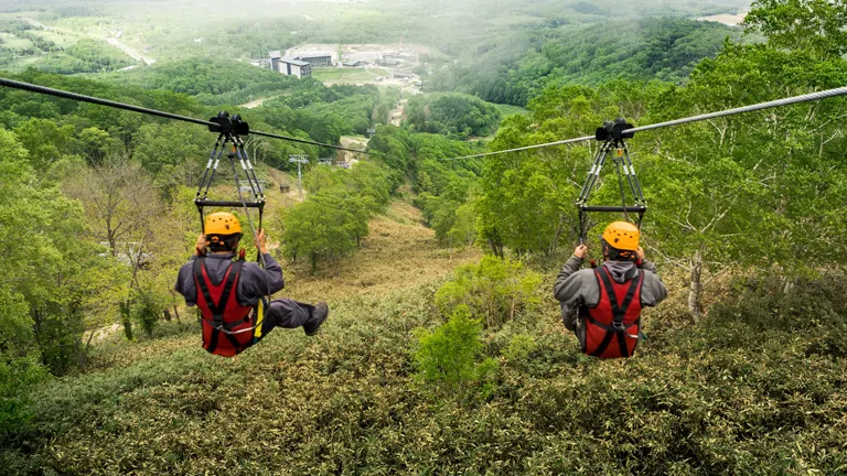 Two people wearing safety gear are zip-lining over a lush green forest, with a clear view of the valley and buildings in the distance.

