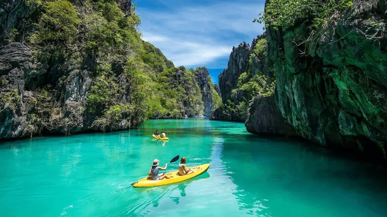 Two kayaks navigate a stunning turquoise lagoon flanked by towering limestone cliffs under a clear blue sky.

