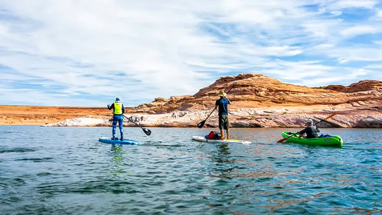 Two people on paddleboards and one in a kayak traverse the calm waters of a lake, with striking layered rock formations in the background.

