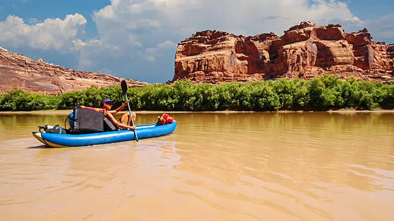 A person in a blue raft on a muddy river, with dramatic red rock formations in the background under a bright, cloudless sky.
