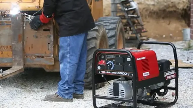 A worker in jeans and a black jacket is welding with a Lincoln Electric Bulldog 5500 AC Welder stationed on gravel ground.