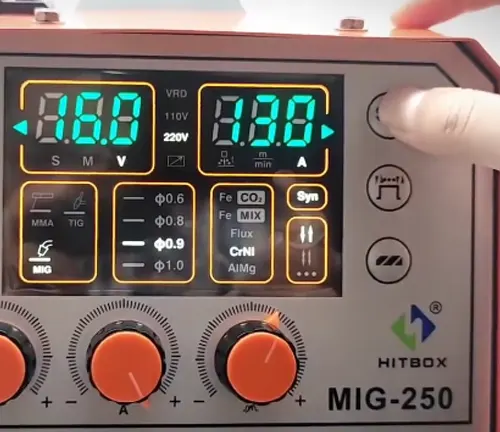 A finger adjusting the settings on the digital control panel of a HITBOX MIG-250 MIG welder with illuminated orange displays.