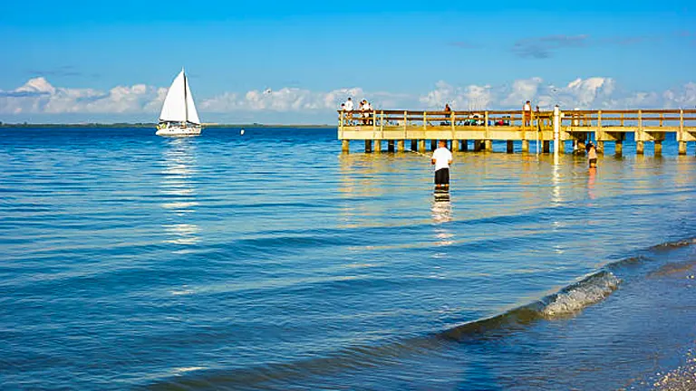 A serene beach scene with a person wading near the shore, a sailboat gliding by in the distance, and a wooden pier with visitors enjoying the view under a clear blue sky.
