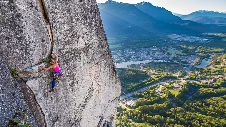A female rock climber in pink scaling a large vertical granite face with a panoramic view of a valley, river, and mountains in the background.
