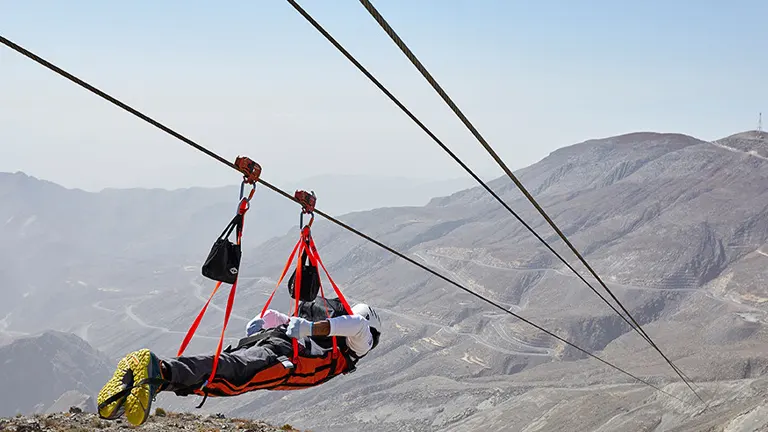 A person in a red and black harness is zip-lining against a backdrop of arid, rocky mountains under a clear sky.

