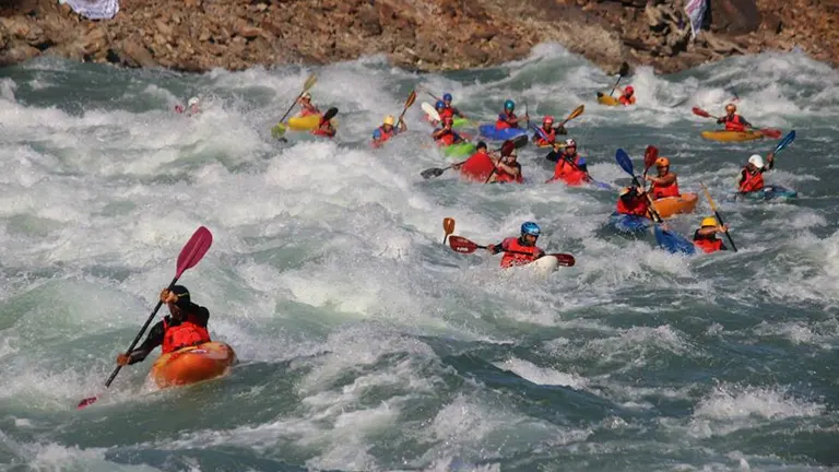 A group of kayakers navigates tumultuous white water rapids, their colorful kayaks and paddles contrasting against the frothy waves.

