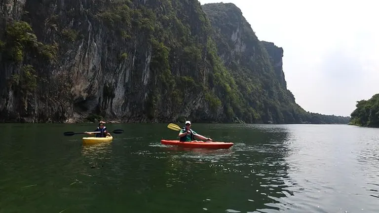 Two people kayaking in calm waters near towering limestone cliffs on an overcast day.

