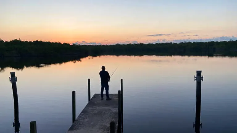 A lone fisherman stands at the end of a tranquil dock during sunrise, with calm waters and a hint of dawn's early light on the horizon, bordered by a serene treeline.

