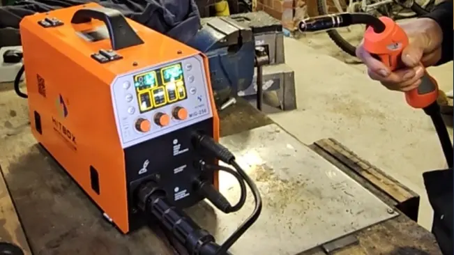 An orange HITBOX MIG200II MIG welder with digital controls on a workshop bench, with a person holding the welding gun nearby.
