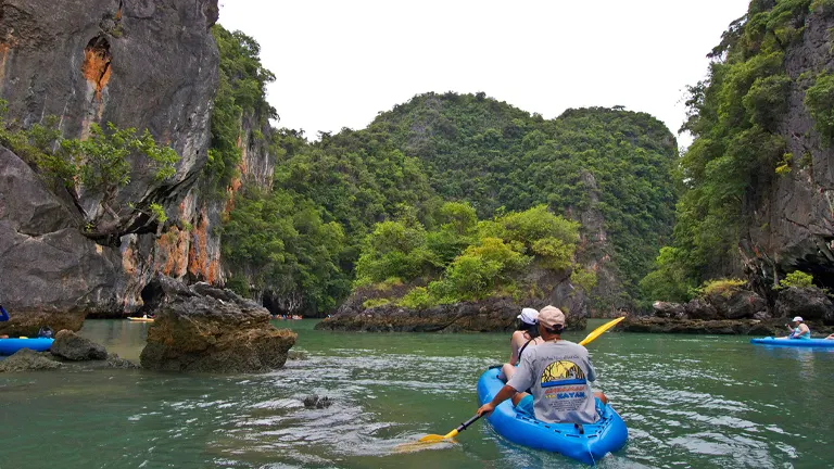 Kayakers paddle in inflatable kayaks through a tranquil water passage surrounded by towering, vegetated limestone cliffs.

