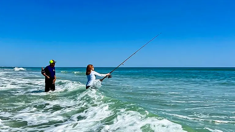 Two people stand in the surf, fishing in the bright blue sea, waves lapping around them under a clear sky, enjoying a sunny day on the coast.
