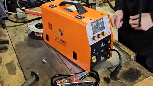 An orange HITBOX MIG200II MIG welder on a workshop table with a digital display, surrounded by various tools and a person's hands working in the background.