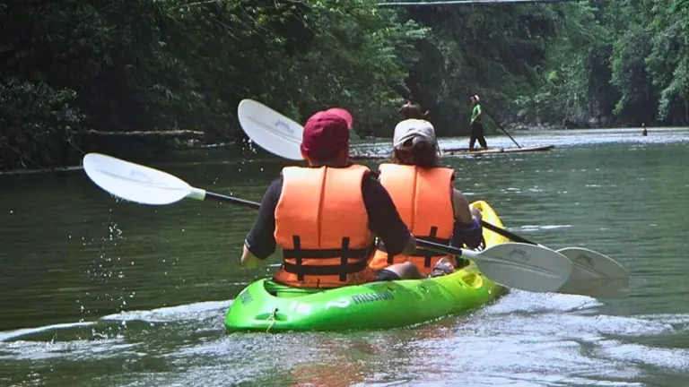Two individuals in life jackets paddling a green kayak on a river flanked by lush forest, with a stand-up paddleboarder visible in the distance.

