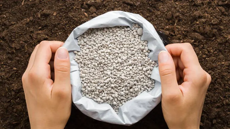 Hands holding open a bag of gray granular fertilizer over rich, dark soil, ready for application.
