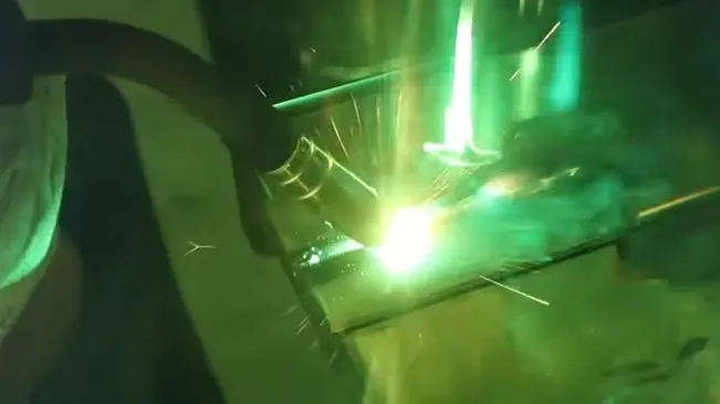 Bright welding arc and sparks captured in a greenish tint, with a welder's hand guiding the process.