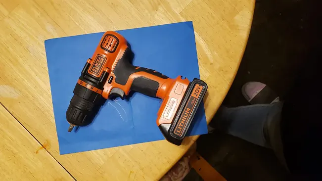 Orange and black BLACK+DECKER cordless drill on blue paper atop a wooden table, with a person's leg visible