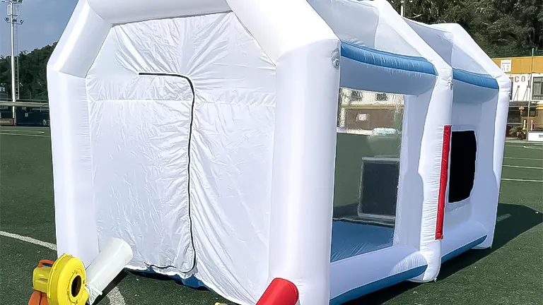 A white and blue inflatable paint booth with a covered entrance and a yellow blower, positioned on an athletic field