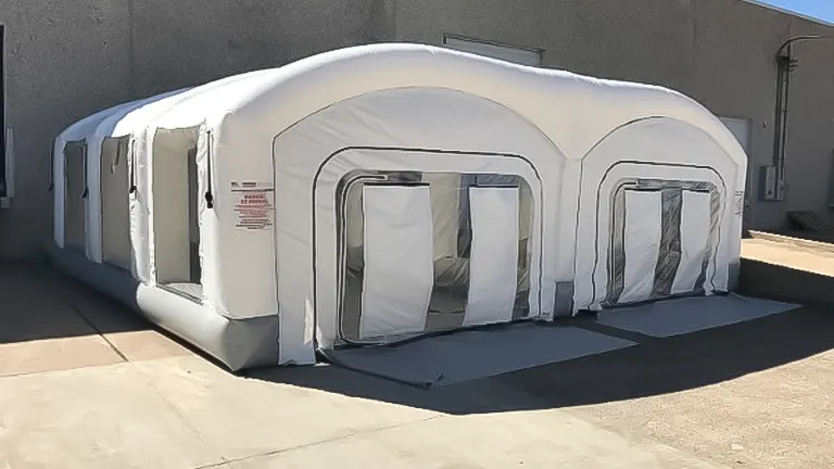 A large, white, inflatable portable paint booth with multiple entrances, positioned on a concrete surface adjacent to a building