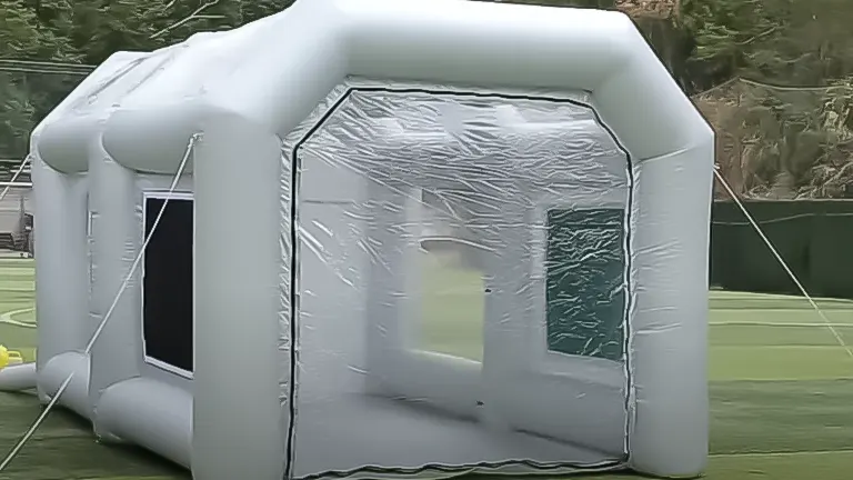 A gray, inflatable paint booth with clear plastic windows, erected on a grassy field with ropes securing it in place