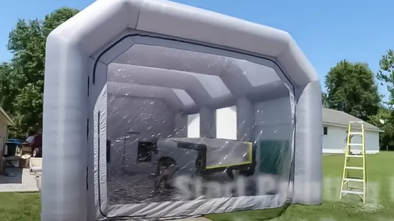 Inflatable gray paint booth with a clear viewing panel, housing a partially covered vehicle, set up in a grassy outdoor area