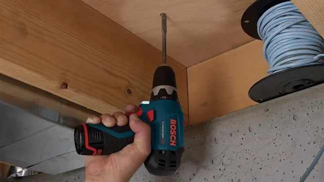 A person using a Bosch PS31-2A 12V Max Drill/Driver Kit to drive a screw into a wooden surface, with a spool of cable in the background.