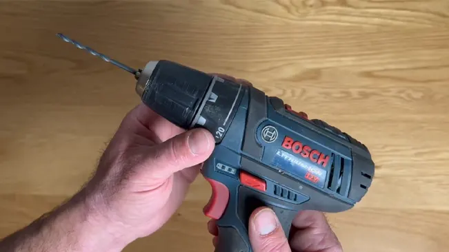 Hand holding a Bosch PS31-2A drill with a bit, against a wooden floor background.