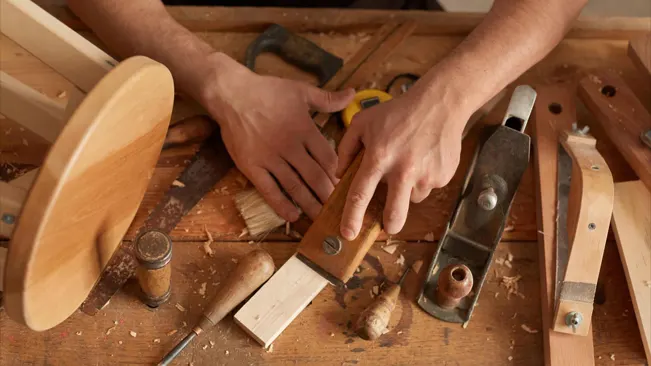 Carpenter's hands using chisel and plane among woodworking tools on a workbench.