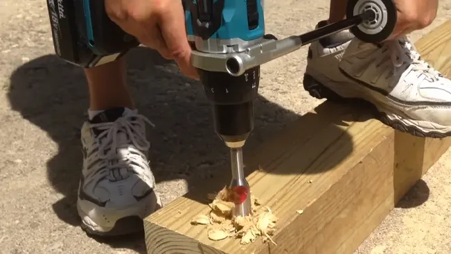 Person using a Makita XPH07Z drill with a large auger bit on a piece of wood outdoors.