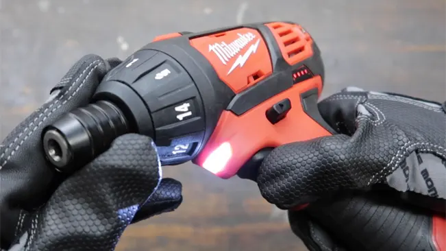 Gloved hands holding a Milwaukee M12 cordless screwdriver with lit LED light