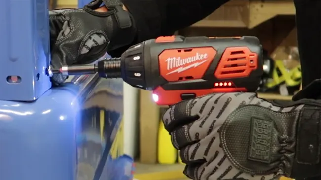Hands in gloves using Milwaukee M12 cordless screwdriver with LED light on blue metal.