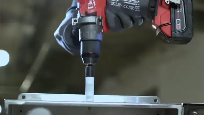 A red and black Milwaukee cordless hammer drill being operated by a person in gloves to drive a bit into metal