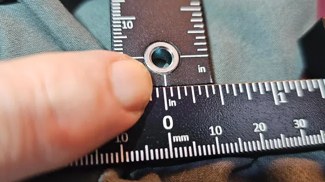 Close-up of a metal grommet being measured with a ruler on teal and brown fabric.