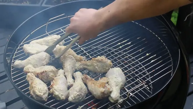 Person grilling chicken pieces on an outdoor barbecue grill.