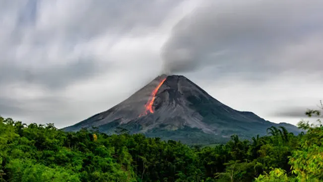 An active volcano with a lava flow, set against a forested landscape, illustrating volcanic activity as a natural cause of wildfires.