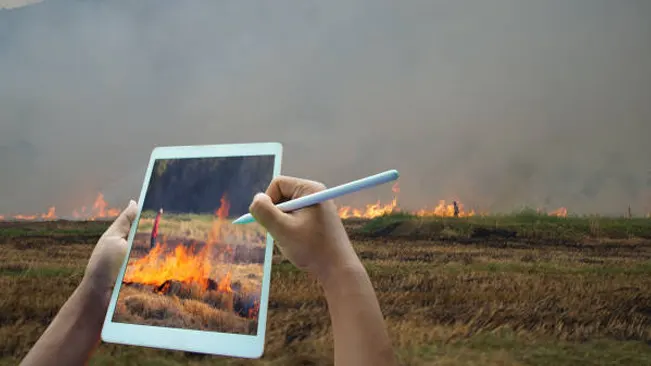 Hands holding a tablet and stylus, digitally documenting a forest fire in the background, signifying the analysis of fire behavior.