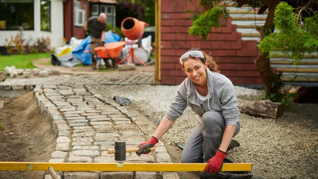 A smiling woman laying cobblestone for a garden path, exemplifying preparation and mitigation efforts in landscaping.