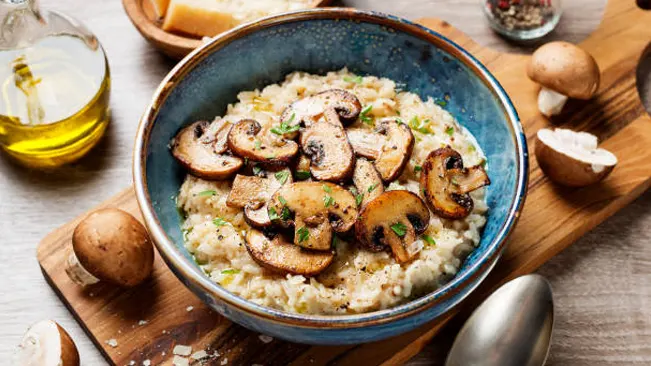 A bowl of creamy risotto topped with sautéed sliced mushrooms, on a wooden board with whole mushrooms and olive oil in the background.
