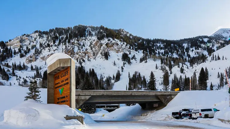 A winter scene showing an entrance with a sign for "The Cliff" lodge or area, with a snow-covered road leading under a bridge, surrounded by vehicles and a mountainous backdrop under a clear sky.