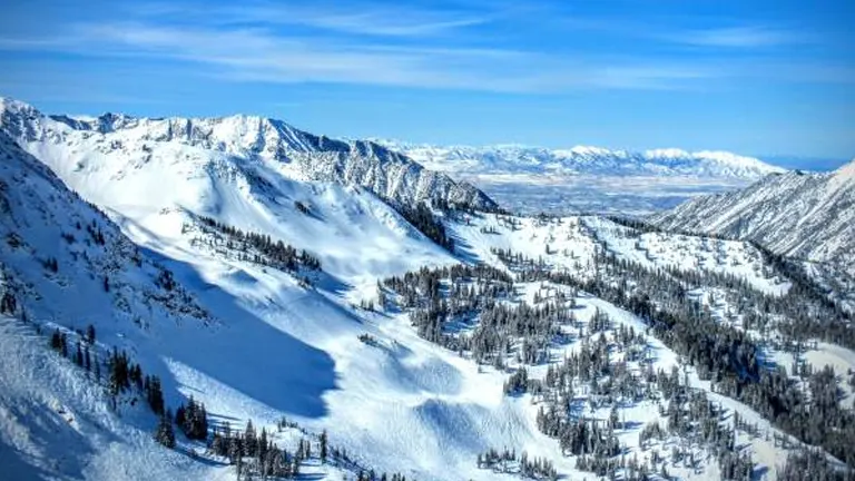 A vast view of a snowy mountain valley with rugged peaks under a bright blue sky, featuring scattered evergreens and traces of ski runs, with distant views that hint at a sprawling, flat landscape beyond the mountains.