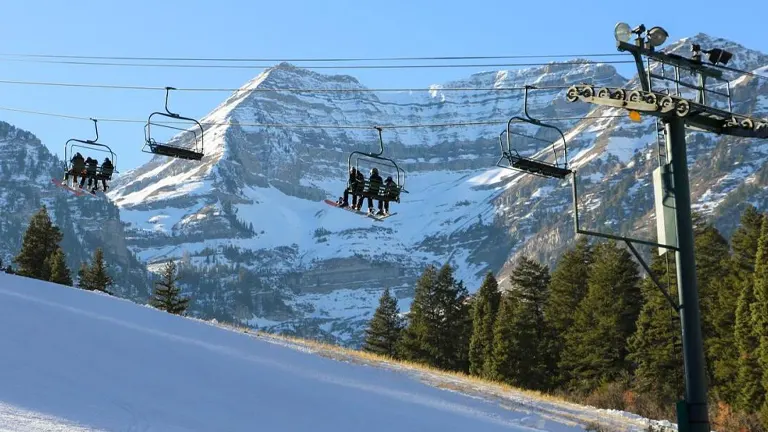 Skiers are riding up a chairlift against the backdrop of a rugged, snow-covered mountain, with clear blue skies and evergreen trees dotting the landscape.